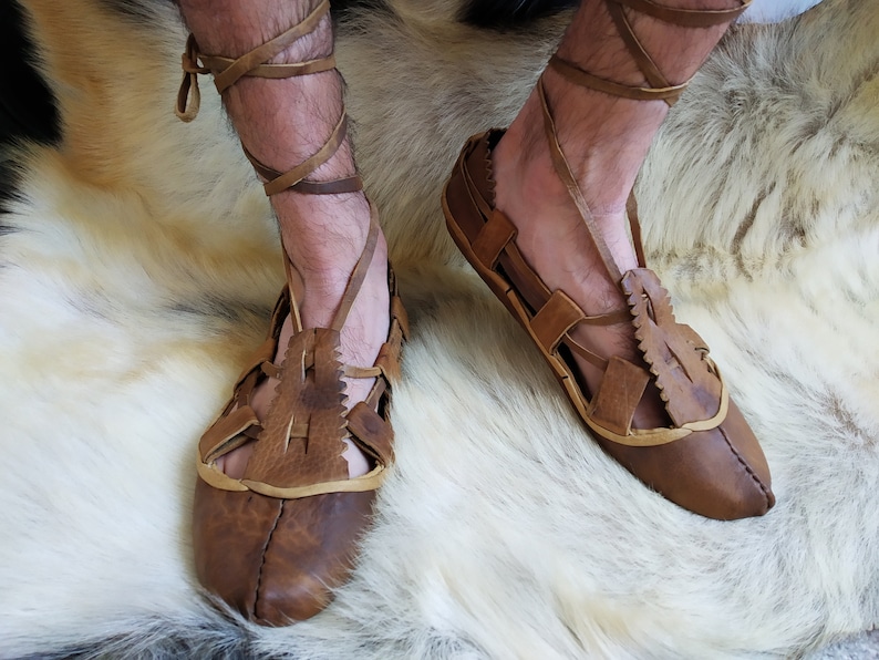 May include: A pair of brown leather sandals with laces that wrap around the ankles. The sandals have a simple design with cutouts and are worn on a white furry surface.