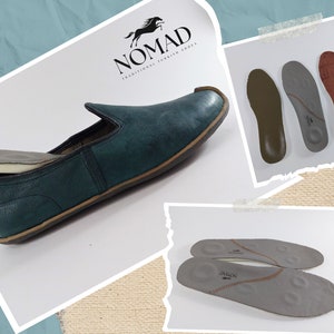 May include: A pair of green leather slip-on shoes with a brown sole. The shoes are shown with a pair of gray insoles. The insoles have the text "Nomad" printed on them. The shoes are made by Nomad, a company that makes traditional Turkish shoes.