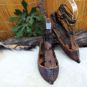 May include: A pair of brown leather sandals with black laces. The sandals have a pointed toe and are decorated with leather straps and stitching.
