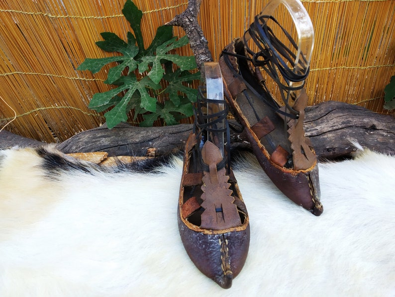 May include: A pair of brown leather sandals with black laces. The sandals have a pointed toe and are decorated with leather straps and stitching.