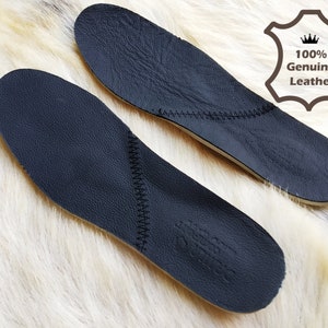 May include: Two black leather insoles with stitching around the edges. The insoles are on a white furry surface. The text "100% Genuine Leather" is visible in the upper right corner of the image.