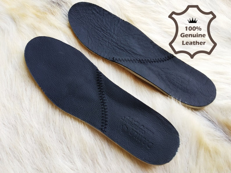 May include: Two black leather insoles with stitching around the edges. The insoles are on a white furry surface. The text "100% Genuine Leather" is visible in the upper right corner of the image.