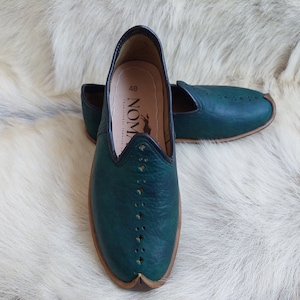 May include: A pair of dark green leather slip-on shoes with a pointed toe and decorative stitching. The shoes have a brown leather trim around the sole and a black leather trim around the opening. The shoes are on a white furry surface. The text "NOM" is visible on the inside of the shoe.