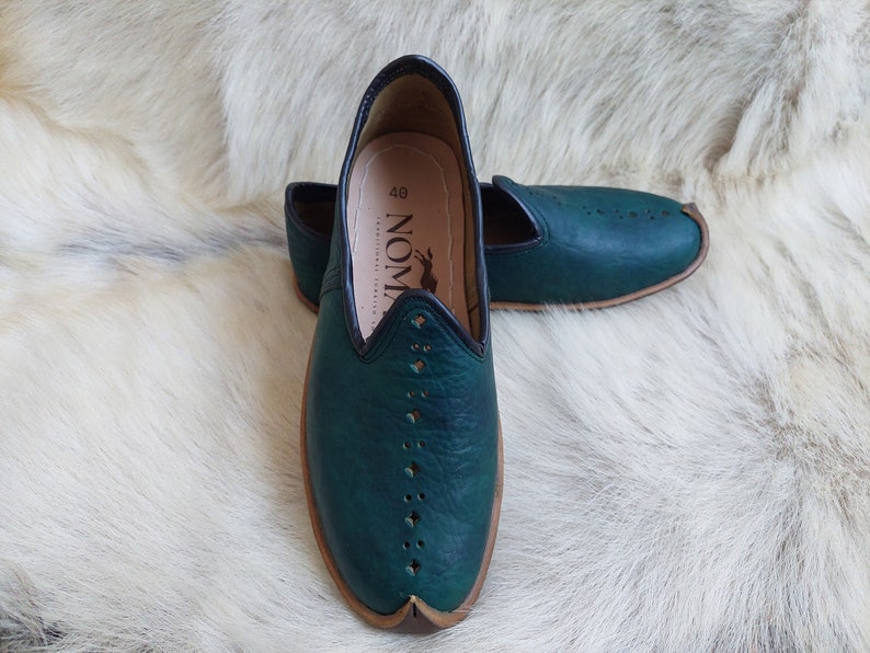 May include: A pair of dark green leather slip-on shoes with a pointed toe and decorative stitching. The shoes have a brown leather trim around the sole and a black leather trim around the opening. The shoes are on a white furry surface. The text "NOM" is visible on the inside of the shoe.