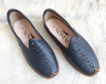 Op de afbeelding: Een paar zwarte leren instapschoenen met een bruine leren rand. De schoenen hebben een geperforeerd ontwerp en de merknaam "NOMAD" staat gedrukt op de binnenzool.