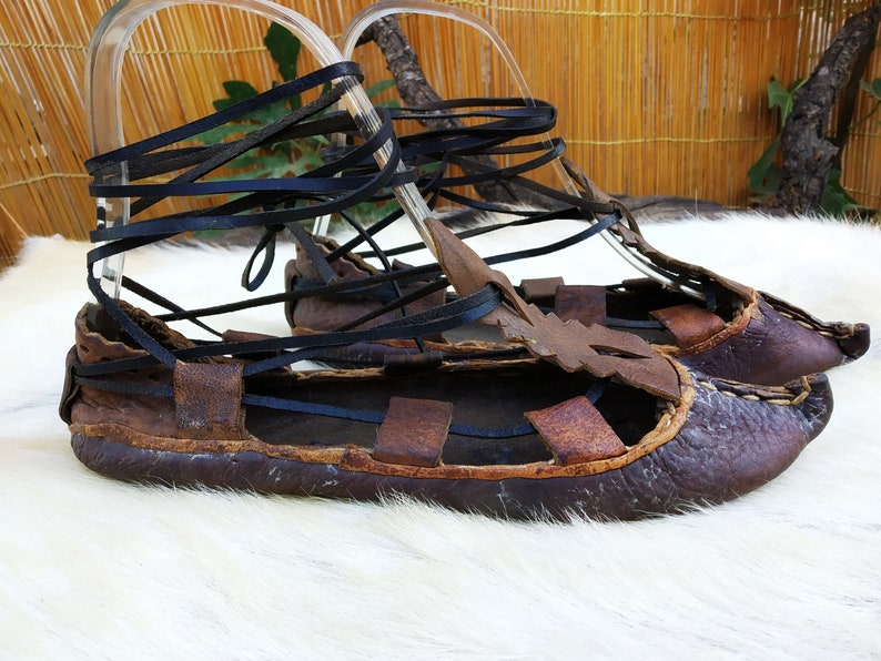 May include: A pair of brown leather sandals with black leather laces. The sandals are tied up the legs and have a pointed toe.