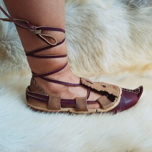 May include: A pair of brown leather sandals with burgundy straps and laces. The sandals have a pointed toe and are tied up the ankle.