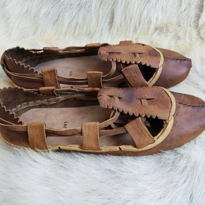 May include: A pair of brown leather sandals with a woven design and a light brown trim. The sandals have a strap across the top of the foot and a strap around the ankle.