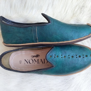 May include: A pair of teal blue leather slip-on shoes with a brown sole. The shoes have a decorative stitching pattern and the insole is labeled "40 NOMAD TRADITIONAL TURKISH SHOES".