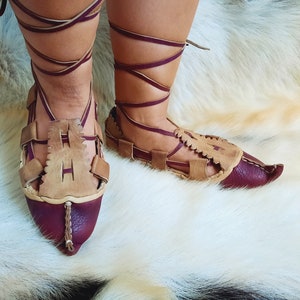May include: A pair of brown leather sandals with burgundy accents and laces. The sandals have a pointed toe and are tied up the leg.