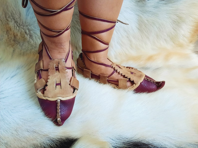 May include: A pair of brown leather sandals with burgundy accents and laces. The sandals have a pointed toe and are tied up the leg.