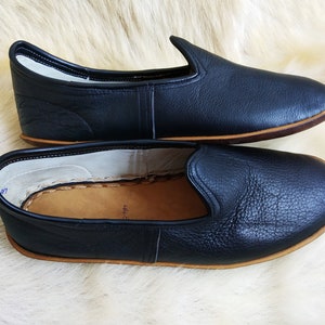 May include: A pair of black leather slippers with a brown leather sole. The slippers are laid out on a white fluffy surface.
