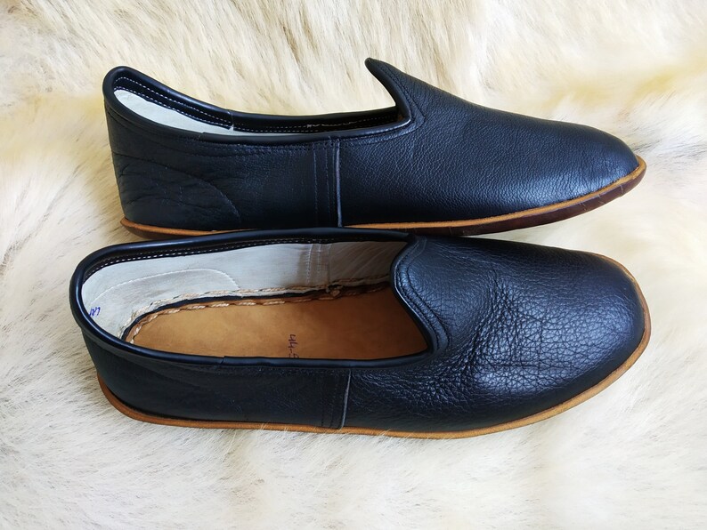 May include: A pair of black leather slippers with a brown leather sole. The slippers are laid out on a white fluffy surface.