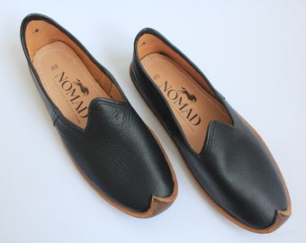 Op de afbeelding: Een paar zwarte leren instapschoenen met een bruine zool. De schoenen hebben een spitse neus en een kleine opening aan de bovenkant. De schoenen hebben het woord "NOMAD" aan de binnenkant gedrukt.