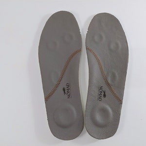 May include: A pair of gray leather insoles with brown stitching and the word "NOMAD" printed on each insole.