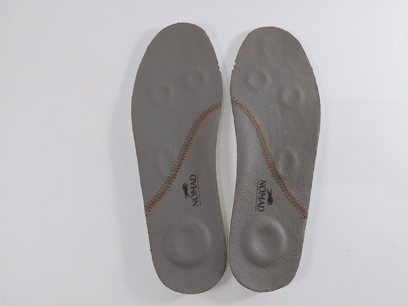 May include: A pair of gray leather insoles with brown stitching and the word "NOMAD" printed on each insole.
