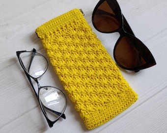May include: A bright yellow, crocheted glasses case with a textured pattern, next to a pair of black-framed eyeglasses and brown-tinted sunglasses. The case is rectangular and has a zipper closure. The sunglasses have a dark tortoiseshell frame.
