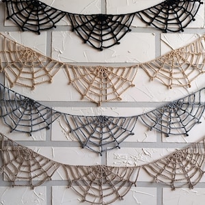 Puede incluir: Cuatro banderines de telaraña de ganchillo en negro, beige, gris y marrón, colgados contra una pared de ladrillos blancos texturizados. Cada banderín presenta un diseño detallado de telaraña, ideal para Halloween.