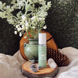 May include: A green glass perfume oil bottle with the text "MOONSEED RITUALS FOREST SPIRIT PERFUME OIL 10 ml" sits on a wooden slice. The bottle is next to a small green vase with white flowers and a clear crystal.