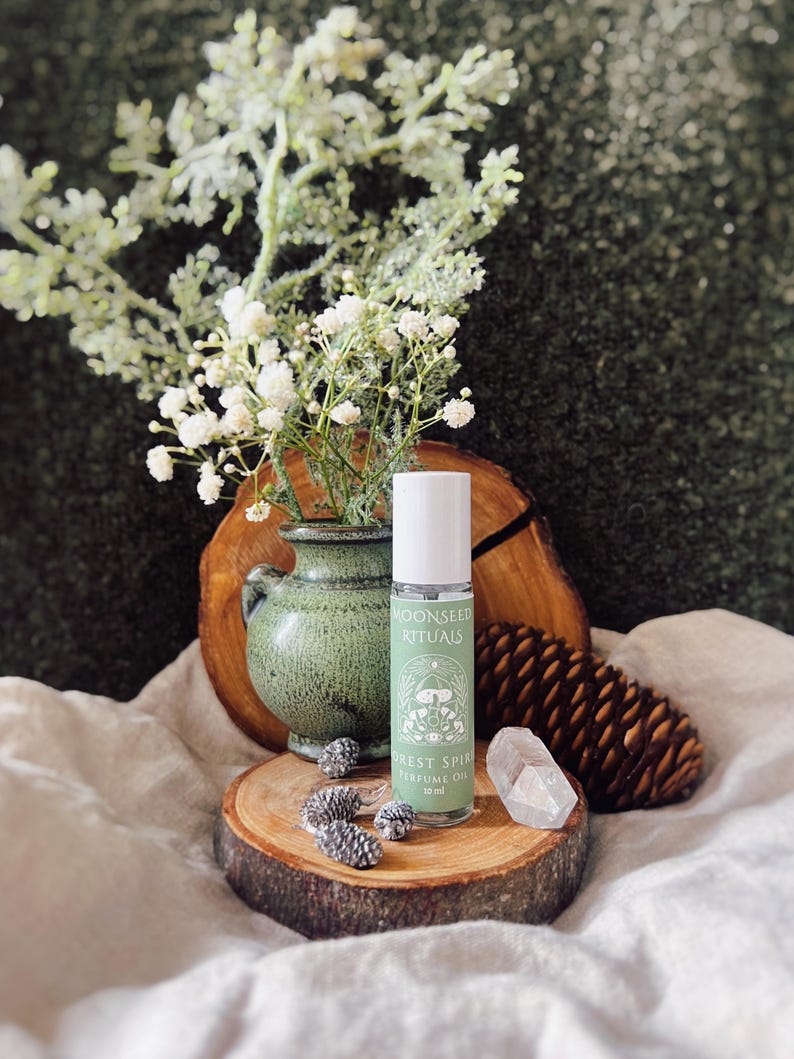 May include: A green glass perfume oil bottle with the text "MOONSEED RITUALS FOREST SPIRIT PERFUME OIL 10 ml" sits on a wooden slice. The bottle is next to a small green vase with white flowers and a clear crystal.