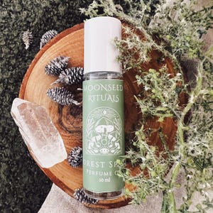 May include: A perfume oil bottle with a white cap and a green label that reads "Moonseed Rituals Forest Spa Perfume Oil 10 ml." The bottle sits on a wooden slice with pinecones, a clear crystal, and greenery.