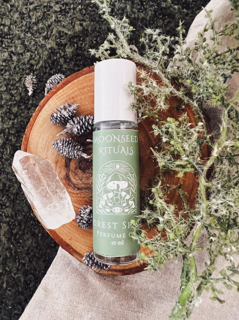 May include: A perfume oil bottle with a white cap and a green label that reads "Moonseed Rituals Forest Spa Perfume Oil 10 ml." The bottle sits on a wooden slice with pinecones, a clear crystal, and greenery.