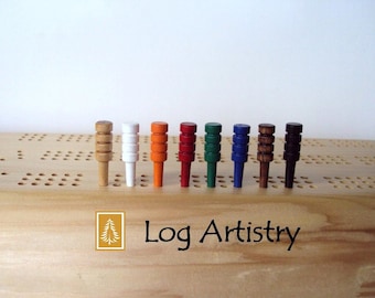May include: A collection of nine cribbage pegs in various colors, including natural wood, white, orange, red, green, blue, and brown. The pegs are arranged on a wooden cribbage board, with the "Log Artistry" logo in the lower left corner.