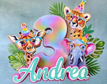May include: A colorful birthday cake topper featuring a number 3 with a rainbow gradient, surrounded by a giraffe, cheetah, and elephant, all wearing glasses and party hats. The name "Andrea" is displayed in a cursive font.