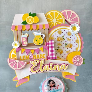 May include: A yellow and pink lemonade stand cake topper with a pink and white gingham number one, the name "Elaina" and the words "Lemonade Stand".