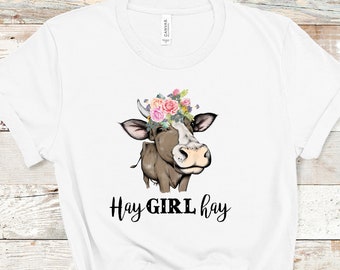 May include: White t-shirt with a brown and white cow wearing a flower crown. The text "Hay GIRL hay" is printed below the cow.