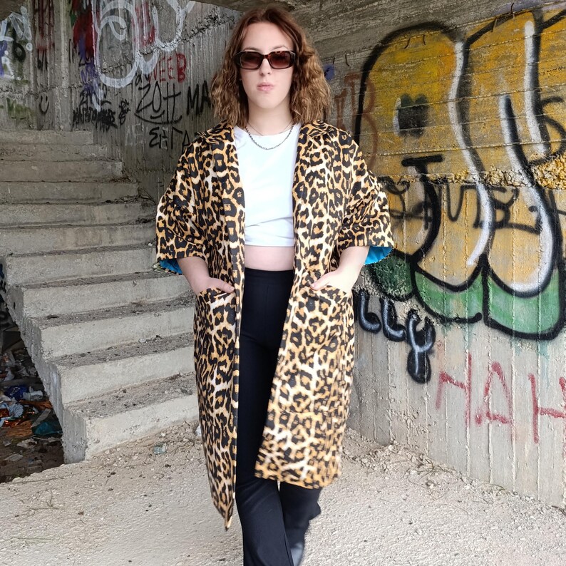 A person with sunglasses and a white crop top wears a leopard print kimono-style coat with teal lining, standing in a concrete underpass with graffiti.