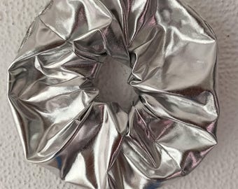 May include: A circular hair scrunchie made of a reflective, metallic silver fabric. The material has a smooth, shiny finish with visible folds and gathers throughout the circular shape, creating a voluminous and textured appearance.