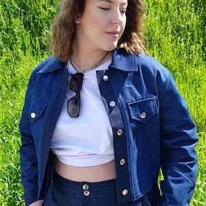 Peut inclure: Une femme portant une veste en jean bleu foncé et un pantalon assorti. La veste est courte et se ferme par des boutons. Le pantalon a une taille haute et une jambe droite.