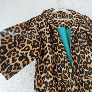 A leopard print kimono-style coat with a teal lining hangs on a white hanger against a white textured wall.