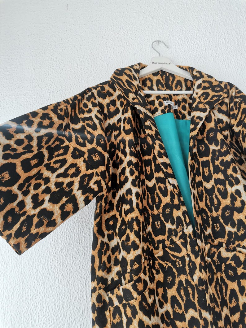 A leopard print kimono-style coat with a teal lining hangs on a white hanger against a white textured wall.