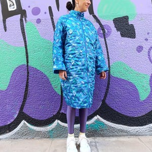 Può includere: Una giacca trapuntata blu con un motivo geometrico. La giacca ha una chiusura a zip e un colletto alto. La modella indossa leggings viola e scarpe da ginnastica bianche.
