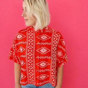 Può includere: Una camicia a maniche corte con bottoni in rosso brillante caratterizzata da un motivo geometrico bianco con rombi e linee a zigzag. La camicia presenta un colletto alla coreana e un pannello verticale di rombi ripetuti lungo l'abbottonatura.