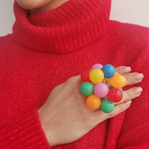 Puede incluir: Un anillo colorido con múltiples bolas pequeñas, redondas y de plástico en varios colores, incluyendo rojo, naranja, amarillo, verde, azul y rosa. El anillo se lleva en un dedo.