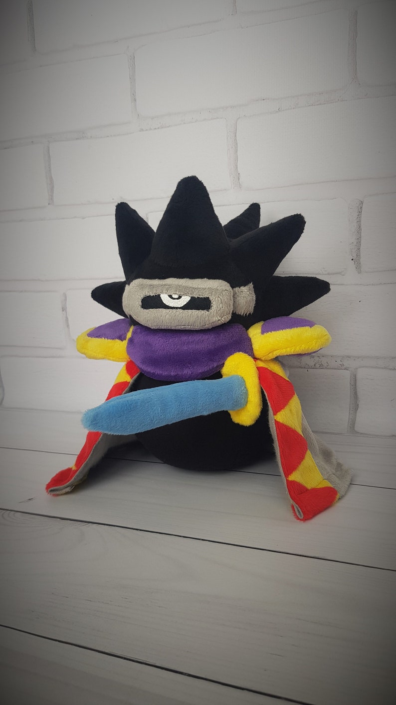 May include: Black plush toy with a spiky head, purple and yellow cape, and a blue sword. The toy has a single white eye.