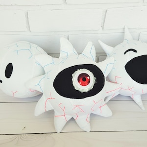 May include: Three white plush toys with different facial expressions. The first toy is a round, white plush with a black face and two black eyes. The second toy is a white plush with a black eye and a red pupil. The third toy is a white plush with a black mouth and a wide, open smile.