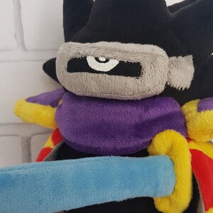 May include: Black plush toy with a purple and yellow cape, a gray mask with a white eye, and a blue sword.