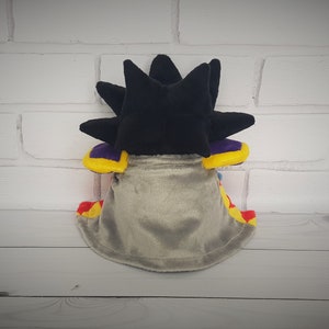 May include: A plush toy with a black spiky head, a gray cape, and yellow, red, and purple accents.