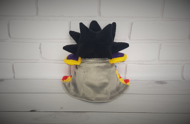 May include: A plush toy with a black spiky head, a gray cape, and yellow, red, and purple accents.