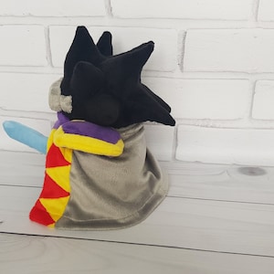 May include: A plush toy with black hair, a purple and yellow cape, and a light blue arm. The toy is standing on a wooden surface.