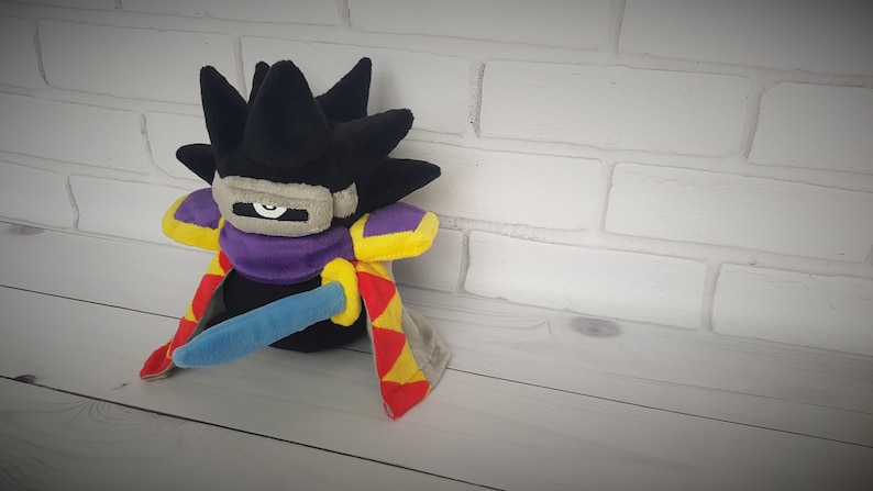 May include: A black plush toy with a purple and yellow cape, a blue sword, and a gray mask over its eyes. The toy is sitting on a white wooden surface.