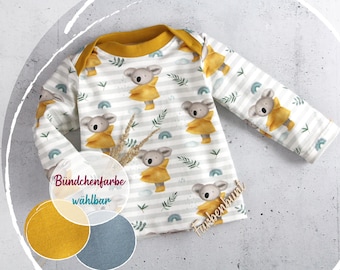 May include: White and gray striped long-sleeved baby shirt with a yellow collar. The shirt features a repeating pattern of koala bears wearing yellow raincoats, with green leaves and blue rainbows. The text "Farbenbunt" is visible.