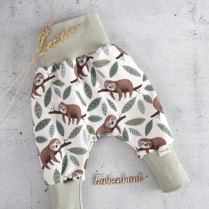 May include: Pants featuring a white background with a repeating pattern of brown sloths on branches and green leaves. The garment has a wide, ribbed sage green waistband and matching ribbed ankle cuffs. A small tag reads Farbenbunt.