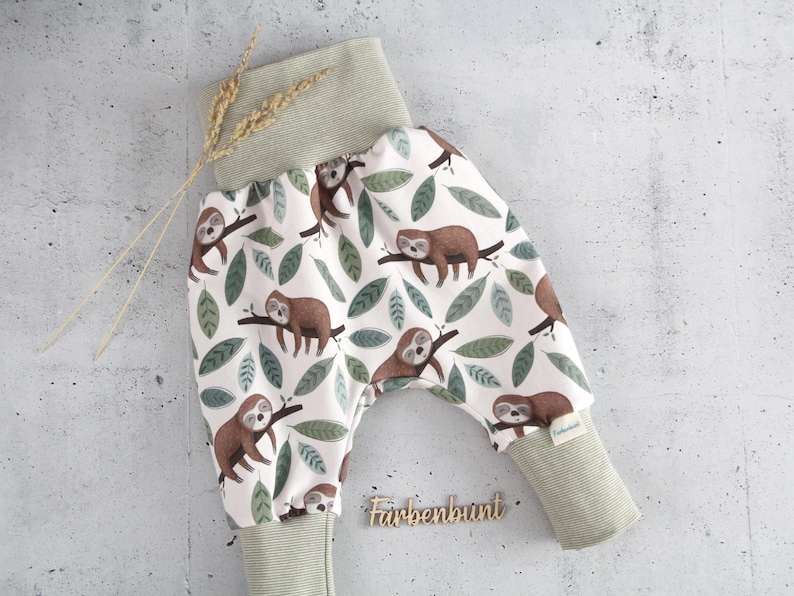 May include: Pants featuring a white background with a repeating pattern of brown sloths on branches and green leaves. The garment has a wide, ribbed sage green waistband and matching ribbed ankle cuffs. A small tag reads Farbenbunt.
