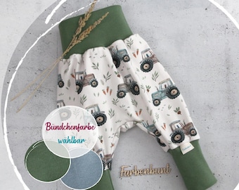 May include: Pants with a cream background featuring a repeating pattern of tractors, carrots, and leaves. The garment has wide, solid olive green elastic cuffs at the waist and ankles. A small tag is visible on the side. Text reads Bündchenfarbe wählbar.