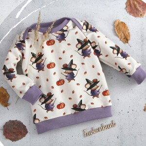 Könnte beinhalten: Ein langärmeliges Pullover-Shirt mit cremefarbenem Hintergrund und einem wiederkehrenden Muster aus braunen Mäusen, die schwarze Hexenhüte und violette Umhänge tragen, durchsetzt mit orangefarbenen Kürbissen und braunen Blättern. Kragen, Bündchen und Saum sind violett.